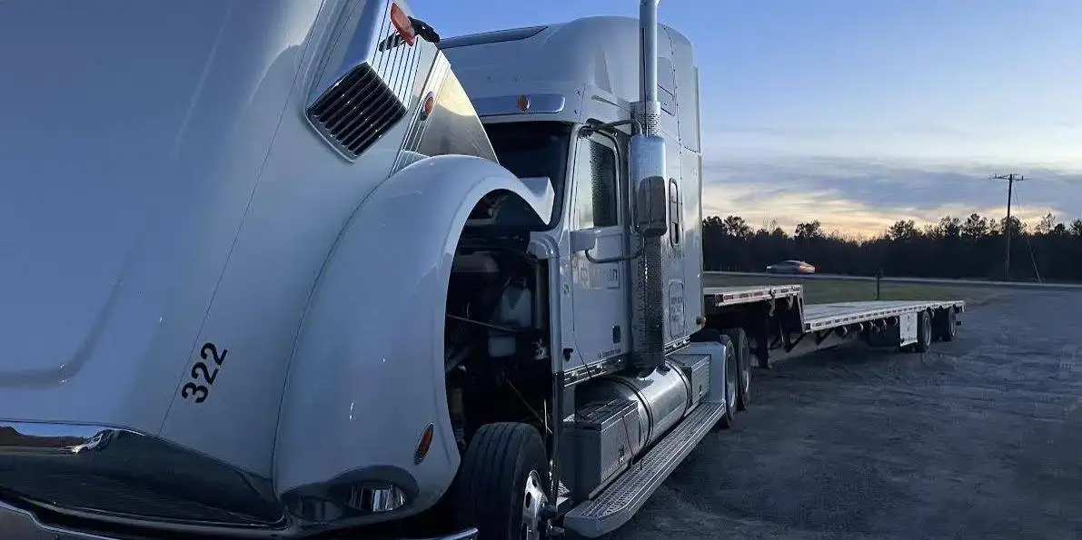 A white Freightliner with its hood tilted open beside its flatbed trailer at dusk