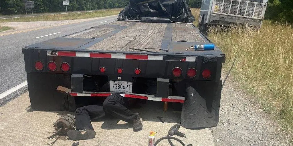A technician working underneath a flatbed trailer on the shoulder of the highway