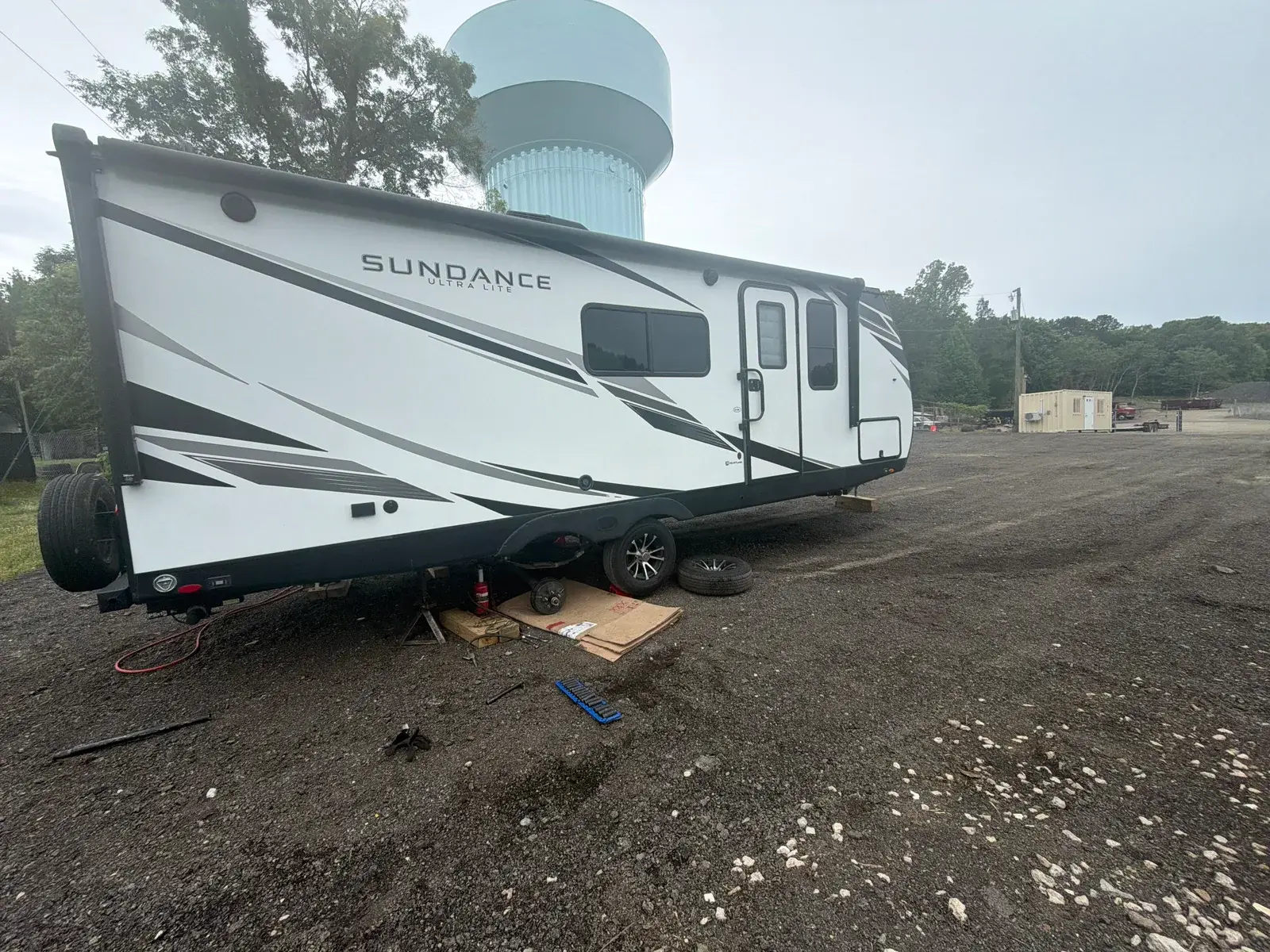 A travel trailer raised on jack stands in the Tri City yard with a road wheel removed and the brake drum exposed, spare wheels and tools on the ground