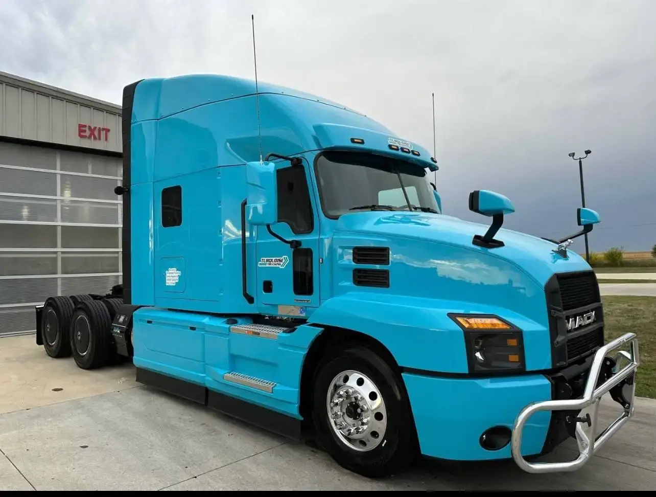 A light blue Mack Anthem sleeper tractor parked outside a workshop bay